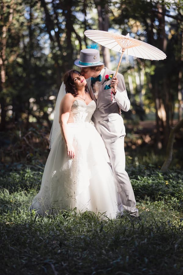 Bride Holly and groom Paul pose outdoors at Kwila Lodge with Paul kissing Holly on the cheek while holding a white parasol.