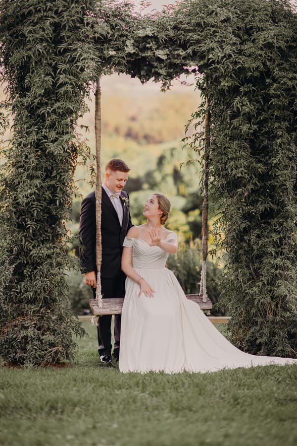 Courtney in a white off-shoulder wedding gown sits on a wooden swing framed by greenery at Tiffany's Maleny, looking up at Liam in a black suit standing beside her.
