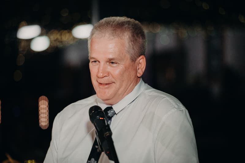 An older man in a white shirt and patterned tie speaks into a microphone at the Preston Peak Winery Functions Centre during the wedding reception.