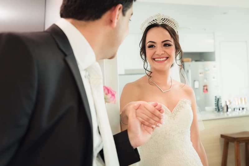The bride Maryam in her wedding dress and tiara holds hands and smiles at the groom Pasha, who is wearing a black suit and white tie, in a bright indoor setting.