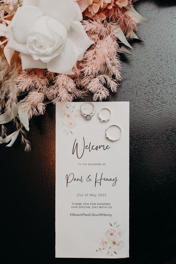 Wedding welcome card for Paul and Henny dated 21st of May 2022 with three rings placed on top and a floral arrangement featuring a white rose and pink foliage on a dark wooden surface.