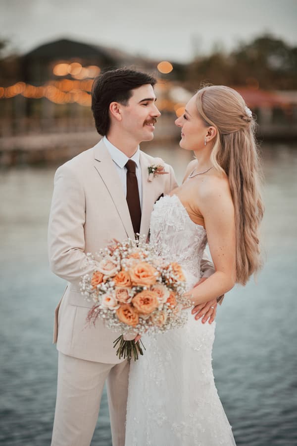 Morgan and Joel pose for couple portraits at Sandstone Point Hotel near the water, with Morgan holding a bouquet of peach and white flowers.