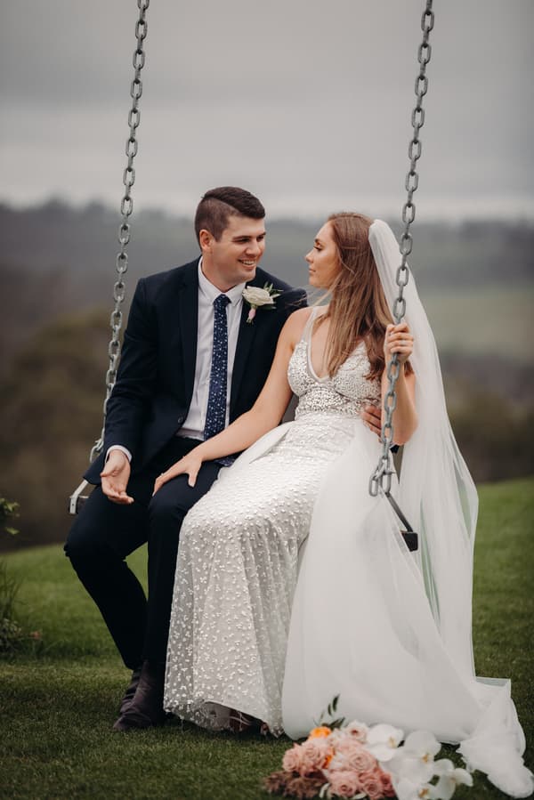 Chelsea and Brent sit together on a swing outdoors at Preston Peak Winery Functions Centre, with Chelsea wearing a white wedding dress and veil and Brent in a dark suit with a boutonniere.