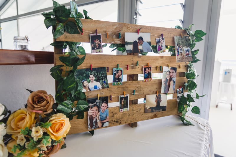 A wooden photo display board decorated with green foliage and holding multiple small photos of a couple and friends, placed on a white tablecloth with a bouquet of yellow and peach roses. Popular wedding seasons 2026 by She Said Yes.