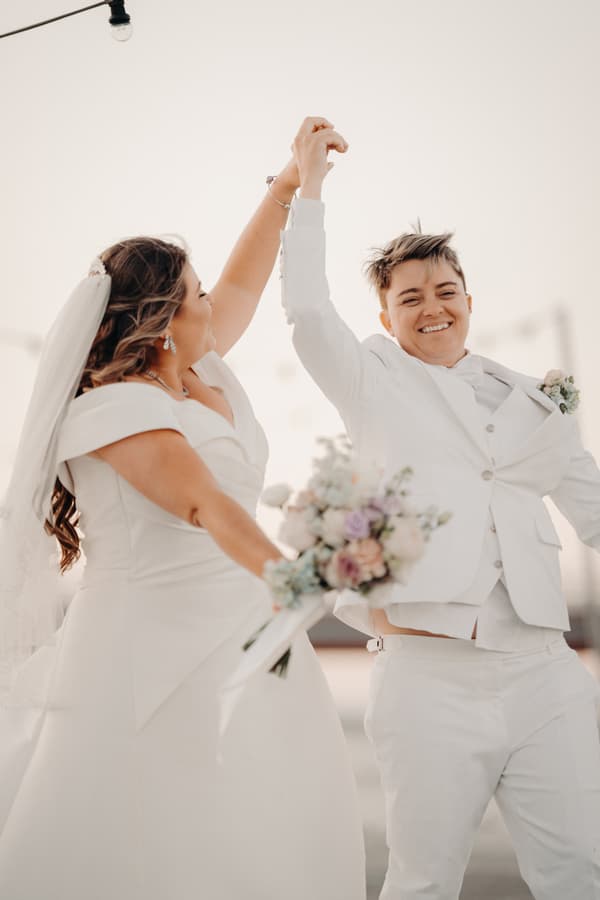 Brooke and Tiffany, both dressed in white wedding attire, hold hands raised in celebration outdoors at Sandstone Point Hotel — The Pavilion.