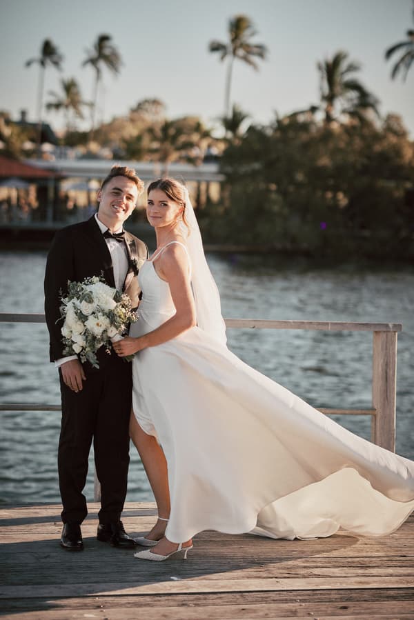 The bride Ashleigh in a white wedding gown holding a bouquet and the groom James in a black tuxedo pose together on a wooden pier at Sandstone Point Hotel with water and palm trees in the background.
