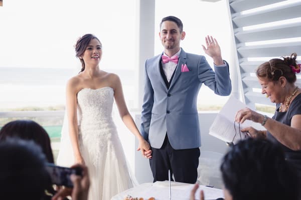 Bride Wing and groom Jason hold hands at the ceremony stage at Bilinga Beach Weddings — The Terrace, with Jason raising his left hand and an officiant holding papers nearby.