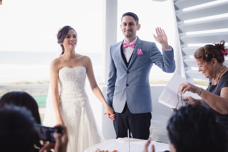 Bride Wing and groom Jason hold hands at the ceremony stage at Bilinga Beach Weddings — The Terrace, with Jason raising his left hand and an officiant holding papers nearby.