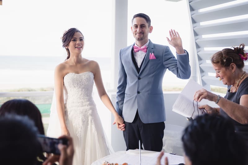 Bride Wing and groom Jason hold hands at the ceremony stage, with Jason raising his left hand and an officiant holding papers nearby. Photo and video packages by She Said Yes.