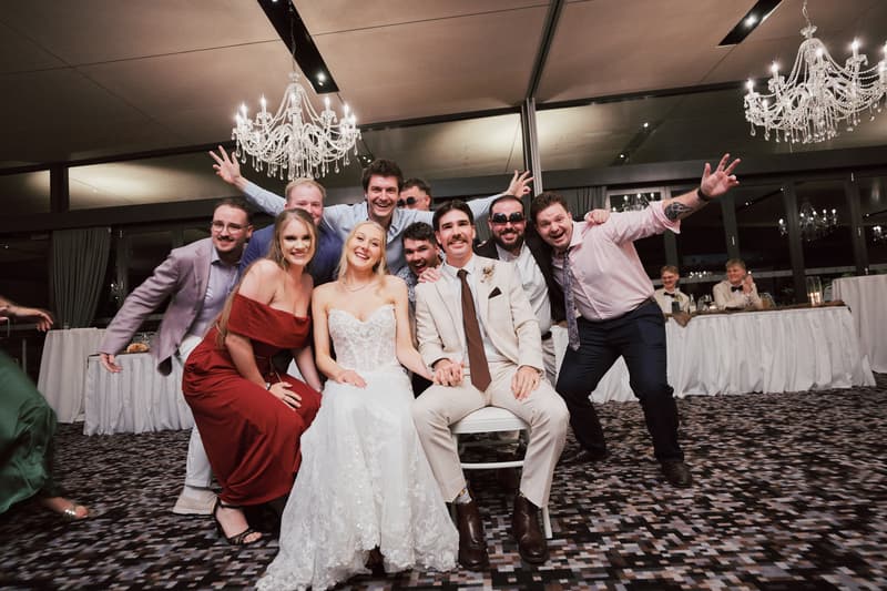 Bride Morgan and groom Joel sit together at Sandstone Point Hotel — The Pumicestone Room reception stage, surrounded by eight guests posing happily behind them.