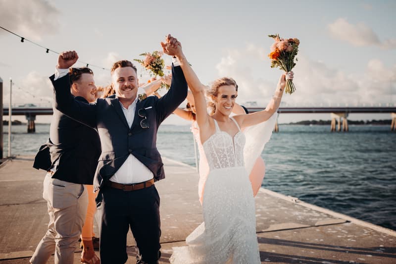 Bride Krystal and groom Brandon celebrate with raised hands on a pier at Sandstone Point Hotel during couple portraits, accompanied by bridesmaids and groomsmen holding bouquets.