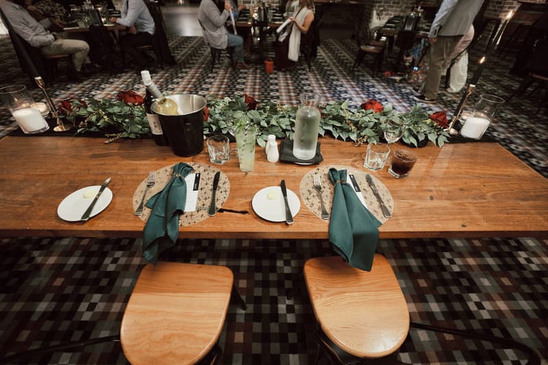 Table set for two at the reception stage with green napkins, plates, cutlery, a bottle of white wine in an ice bucket, a water carafe, glasses, and a floral centerpiece at Sandstone Point Hotel — The Cellar.
