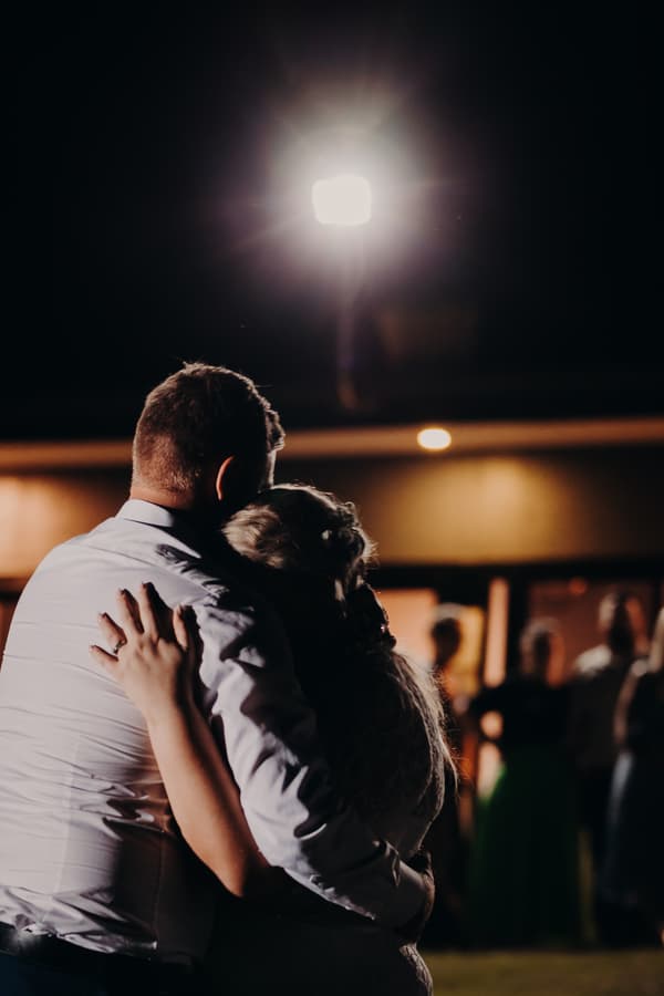 The bride Annie and groom Trent embrace during a dance at the reception stage at Ocean View Estates, with guests blurred in the background and a bright light overhead.