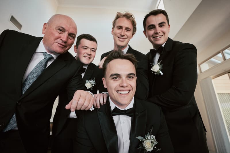 James the groom poses with four men in tuxedos, likely groomsmen and a family member, indoors at Sandstone Point Hotel.