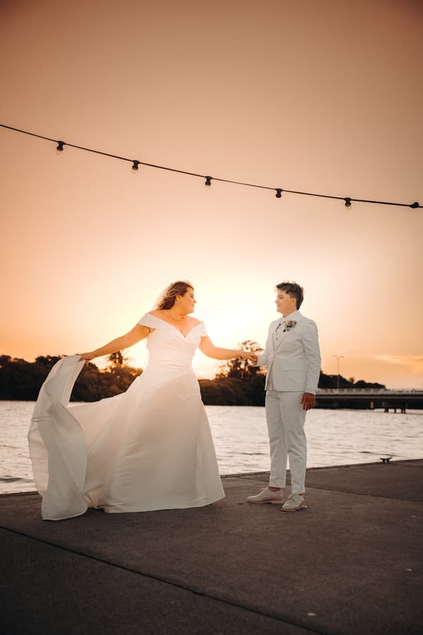 Brooke and Tiffany stand holding hands on a waterfront at sunset, with Brooke wearing an off-shoulder wedding gown and Tiffany in a white suit at Sandstone Point Hotel.