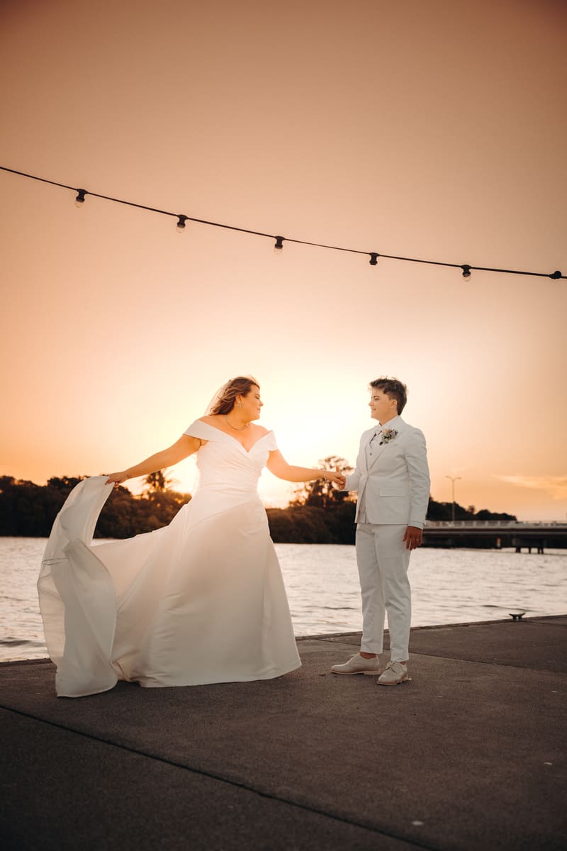 Brooke and Tiffany stand holding hands on a waterfront at sunset, with Brooke wearing an off-shoulder wedding gown and Tiffany in a white suit. Winter wedding venues Brisbane indoor by She Said Yes.