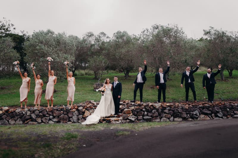 Chelsea the bride and Brent the groom stand together on a stone wall at Preston Peak Winery Functions Centre, flanked by four bridesmaids in matching light-colored dresses holding bouquets raised and four groomsmen in dark suits waving.