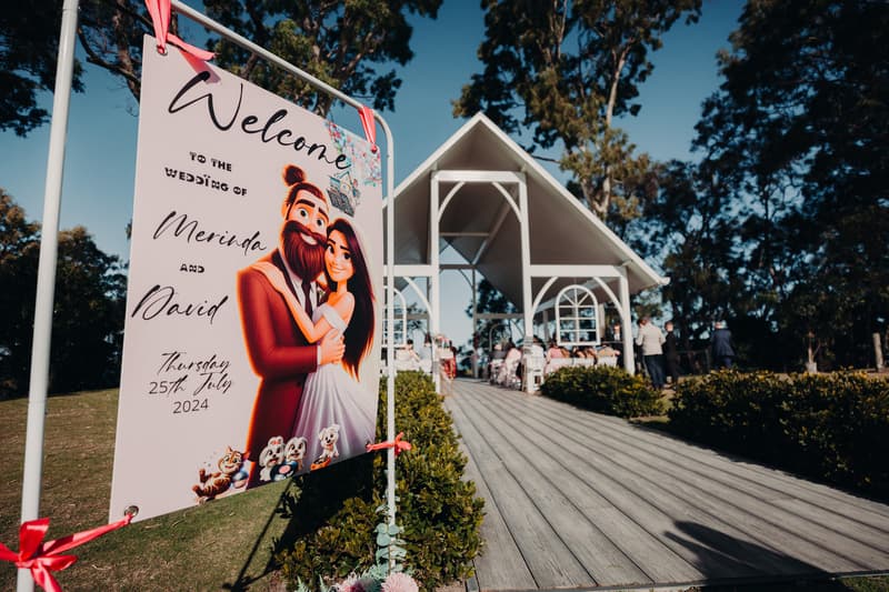 Welcome sign for the wedding of Merinda and David displayed near the ceremony pavilion at Sandstone Point Hotel, with guests seated inside the pavilion in the background.