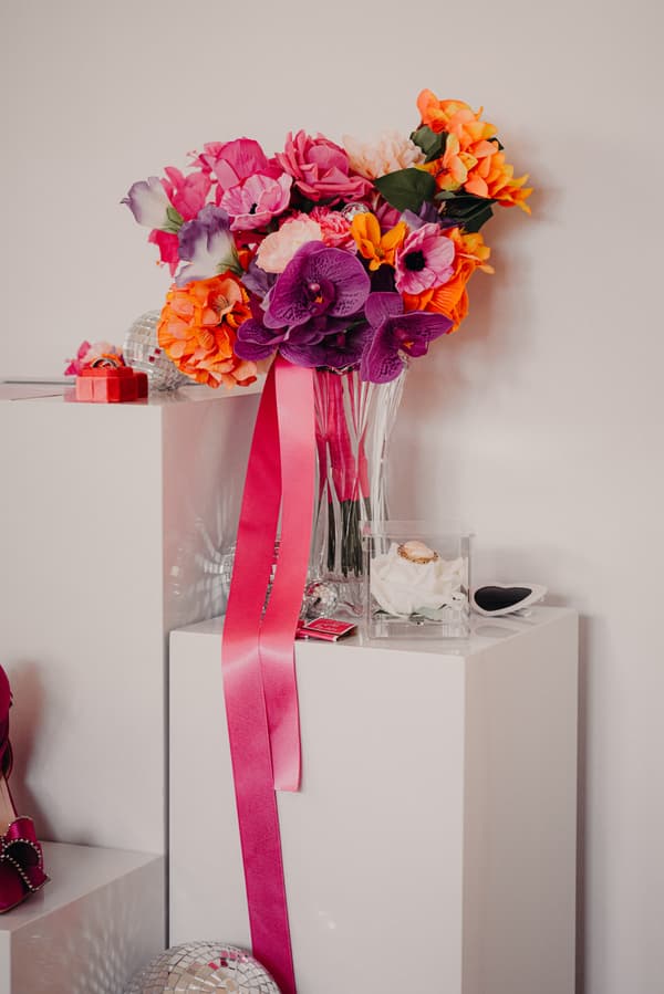 A vibrant bouquet of mixed flowers with pink ribbons in a clear vase, placed on a white pedestal alongside a small clear box containing a white flower and a ring, at Sandstone Point Hotel.