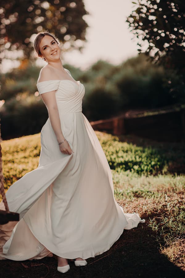 The bride Courtney poses alone outdoors at Tiffany's Maleny, wearing an off-shoulder white wedding gown and holding part of her dress.