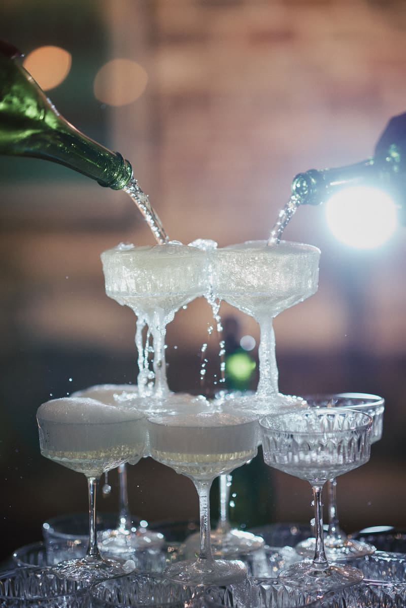 Two bottles pour champagne into a stacked pyramid of coupe glasses. Getting ready photos by She Said Yes.