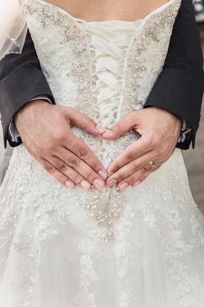 The groom's hands form a heart shape over the bride's lace-up wedding dress back with intricate beadwork and embroidery. Wedding photography add-ons by She Said Yes.