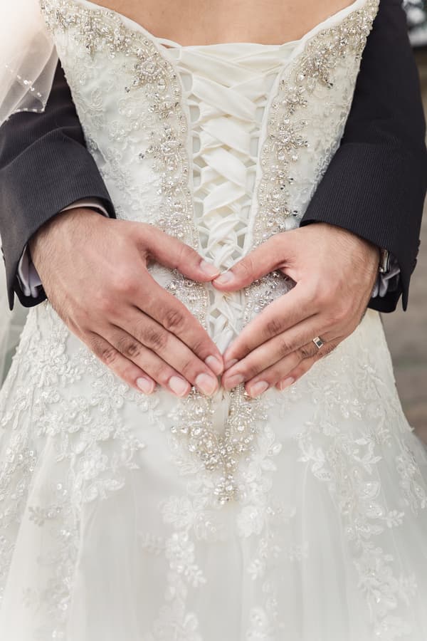 The groom's hands form a heart shape over the bride's lace-up wedding dress back with intricate beadwork and embroidery.