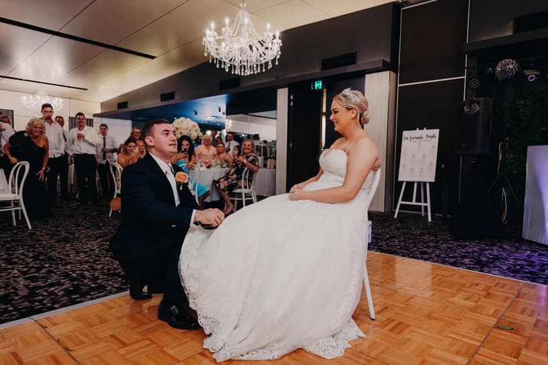The groom kneels on the dance floor in front of the seated bride during the reception at Sandstone Point Hotel — Pumicestone Room, with guests watching and smiling in the background.