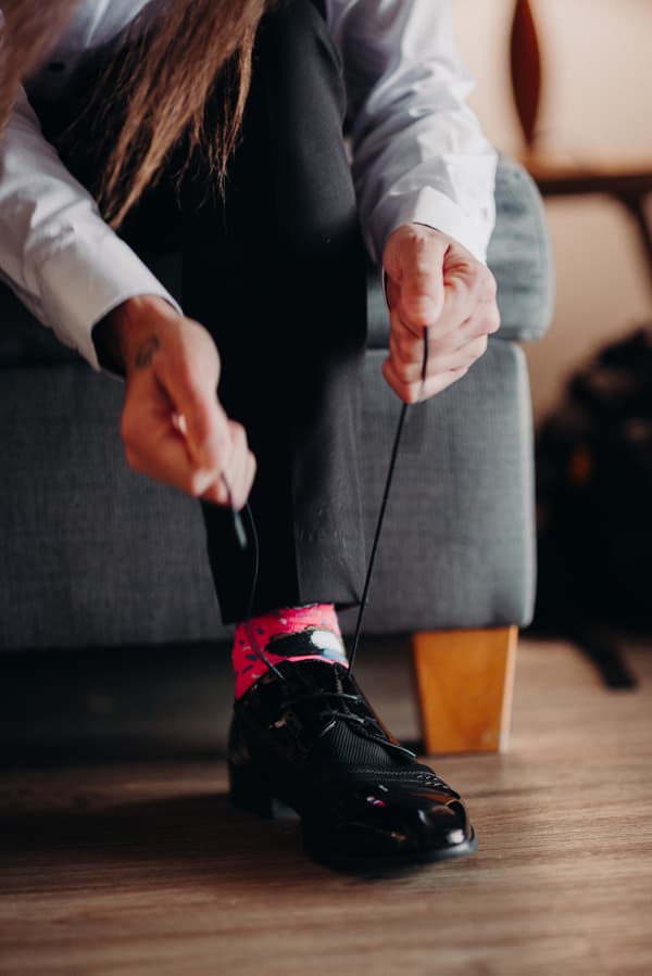 The groom is seated on a gray couch at Sandstone Point Hotel, tying the laces of his black dress shoe, revealing bright pink socks.