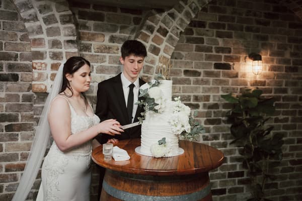 Terion the bride and Tyler the groom cut their wedding cake on a wooden barrel table at Sandstone Point Hotel — The Cellar, with a brick wall and wall light in the background.