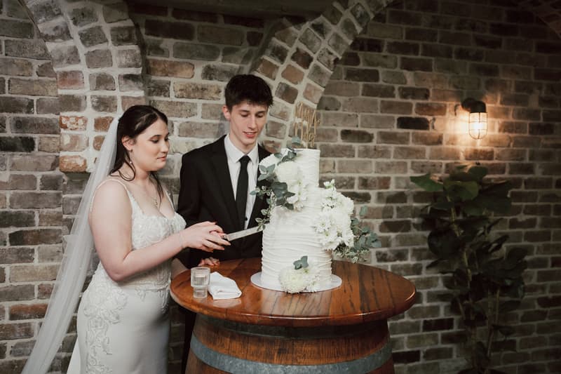 Terion the bride and Tyler the groom cut their wedding cake on a wooden barrel table at Sandstone Point Hotel — The Cellar, with a brick wall and wall light in the background.