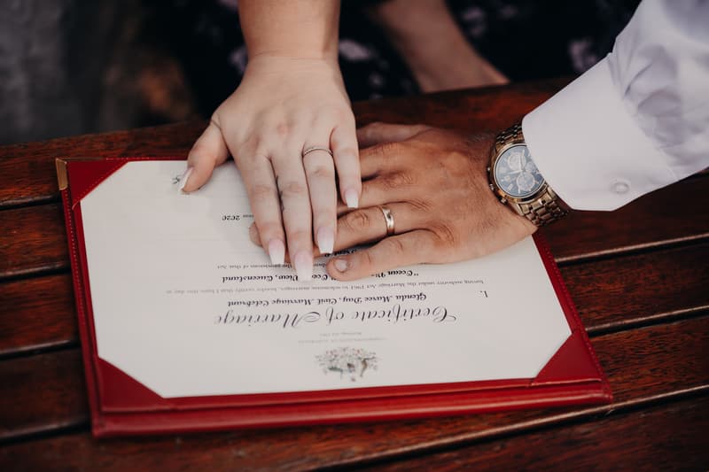 The bride Annie and groom Trent place their hands together over their marriage certificate on a wooden surface at Ocean View Estates.