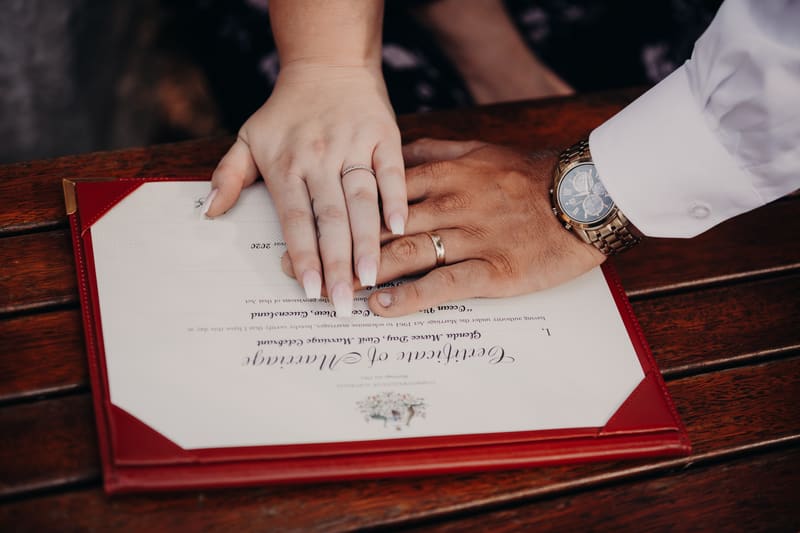 The bride Annie and groom Trent place their hands together over their marriage certificate on a wooden surface. Wedding photography coverage options by She Said Yes.