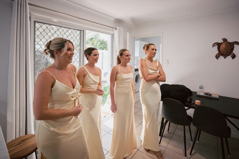 Four bridesmaids in matching cream dresses stand inside a room at Sandstone Point Hotel, near a sliding door with a security screen and white curtains.