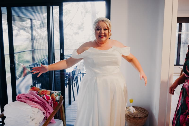 The bride Jacquelyne stands indoors near a sliding glass door at Sandstone Point Hotel, wearing a white off-shoulder wedding dress with arms outstretched.