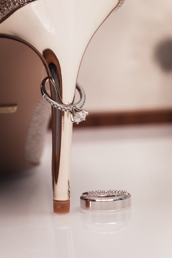 Close-up of a bridal high heel shoe with an engagement ring hanging on the heel and a wedding band placed on the surface nearby.
