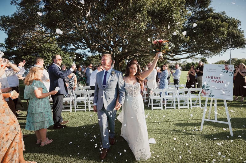 Bride Jheng and groom Dane walk hand in hand down the aisle at Sandstone Point Hotel — The Pavilion after their wedding ceremony, surrounded by guests throwing flower petals.