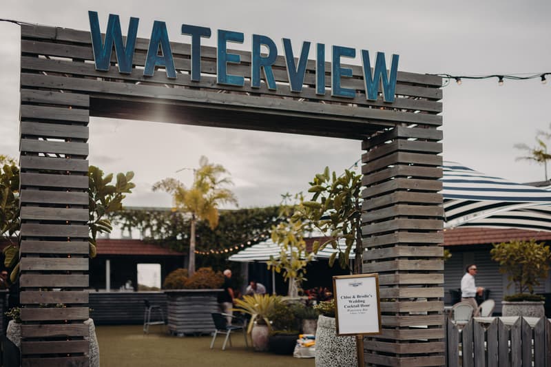 Entrance archway with 'WATERVIEW' sign at Sandstone Point Hotel showing a sign for Chloe and Brodie's wedding cocktail hour with guests and outdoor seating visible in the background.