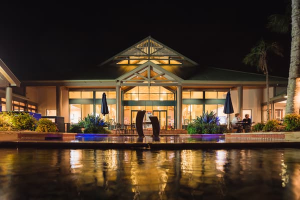 Exterior night view of Novotel Sunshine Coast Resort reception area with illuminated building, outdoor seating, umbrellas, plants, and reflecting pool in the foreground.