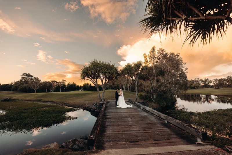 Bride Libby and groom Kyle stand together on a wooden bridge over water at The Tides during a couple portraits session at sunset.