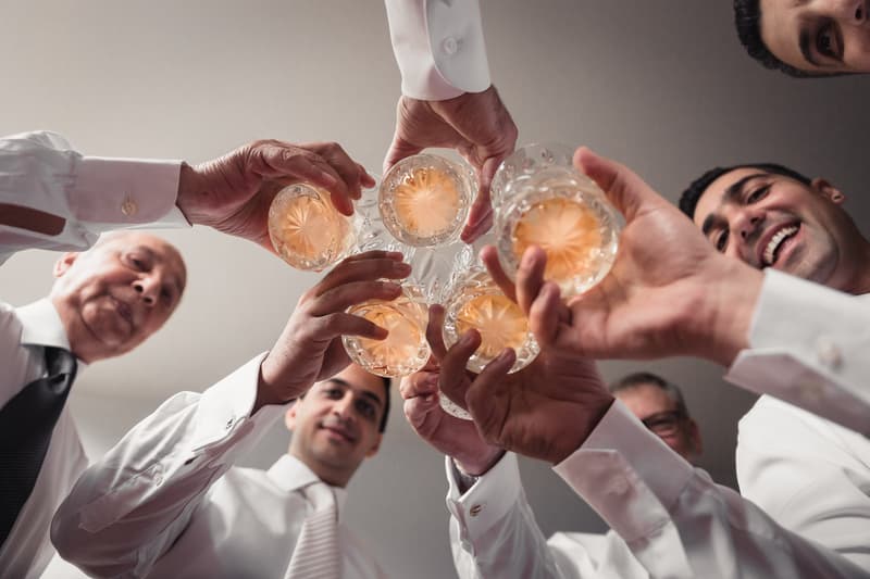 The groom and several men in white shirts and ties raise glasses in a toast, viewed from below against a plain ceiling.