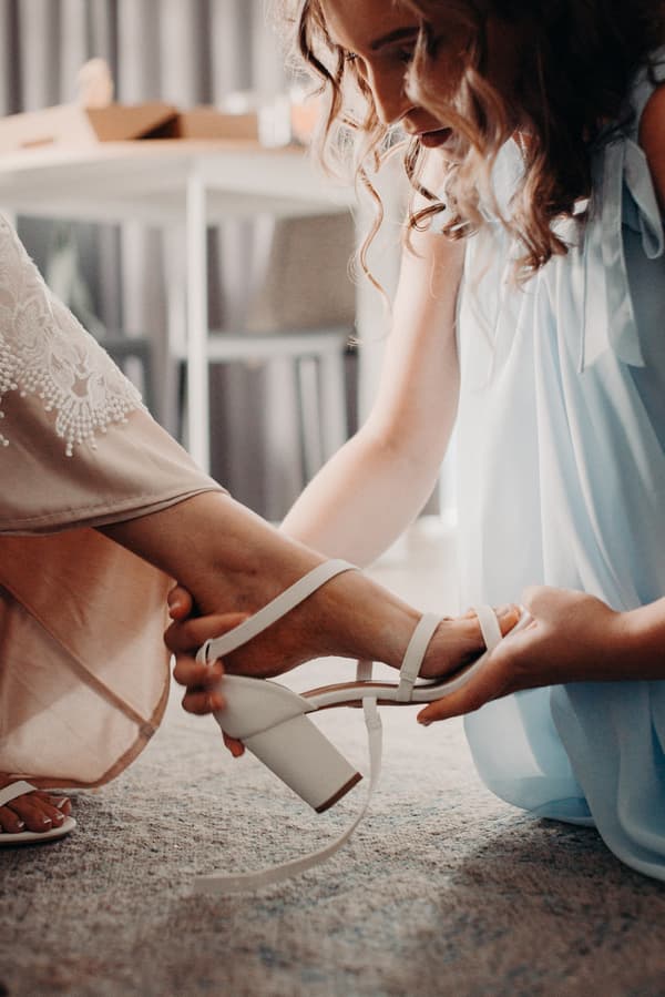 A bridesmaid helps the bride put on a white heeled sandal indoors.