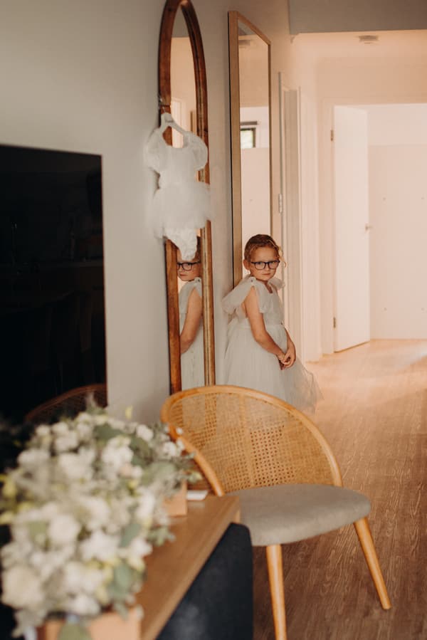 A young flower girl in a white dress and glasses stands near a tall mirror in a hallway at Sandstone Point Hotel, with a small white dress hanging on the mirror and a floral arrangement on a nearby table.