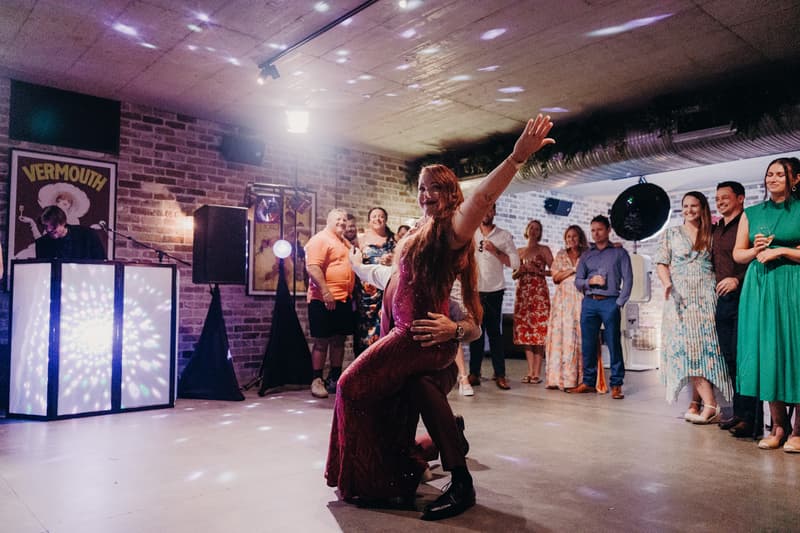 The bride and groom dance on the reception floor at Sandstone Point Hotel — The Cellar while guests watch and a DJ plays music.