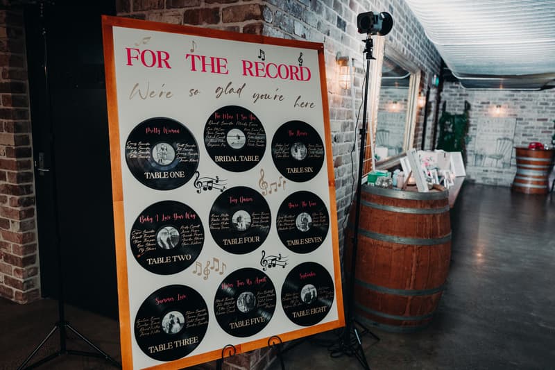 A wedding seating chart titled 'FOR THE RECORD' displayed on an easel at Sandstone Point Hotel — Cellar, featuring table assignments styled as vinyl records with guest names listed under each table.