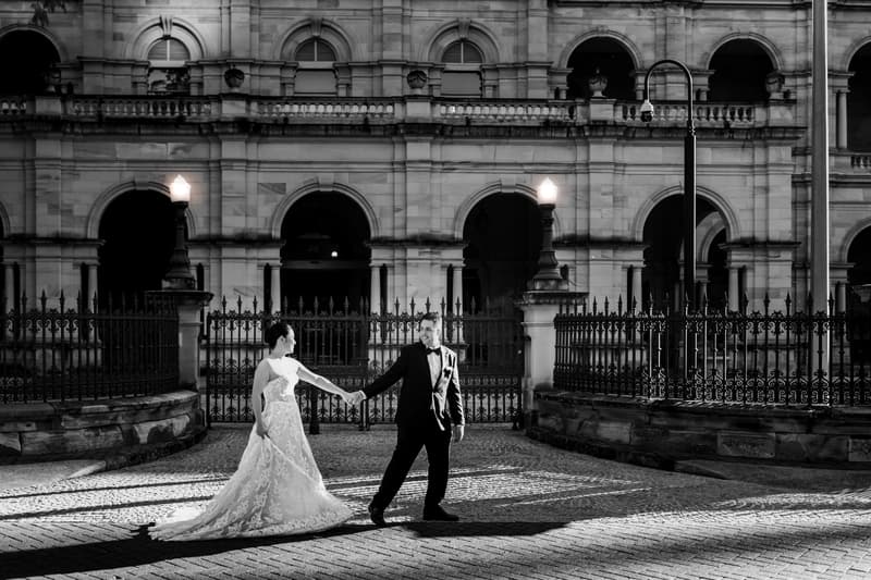 The bride Aria and groom Antony hold hands and face each other in front of the Queensland Parliament building during their couple portraits session.