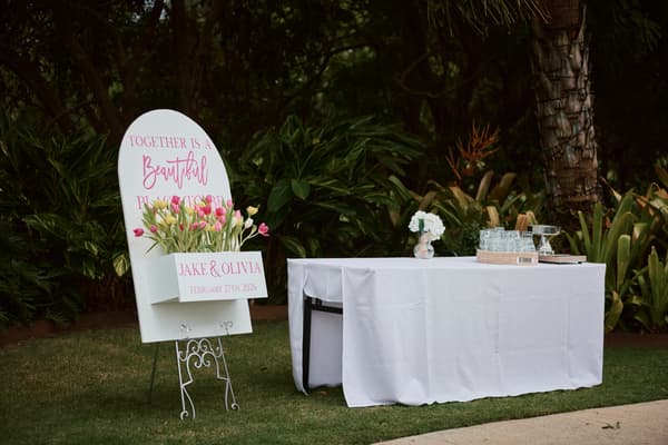 A white sign with pink text and tulips reads 'Together is a Beautiful Place to Be' with the names Jake & Olivia and the date February 27th, 2026, placed next to a white table covered with a cloth holding a small floral arrangement, glasses, and a basket at Eatons Hill Hotel — Lakeside.