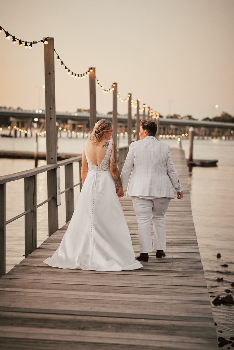 Brides Mandy and Selina walk hand in hand along a wooden pier. Cinematic wedding photos by She Said Yes.