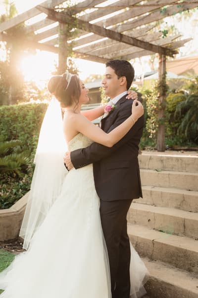 The bride Maryam and groom Pasha embrace outdoors near stone steps under a wooden pergola. Winter wedding Brisbane dress code by She Said Yes.