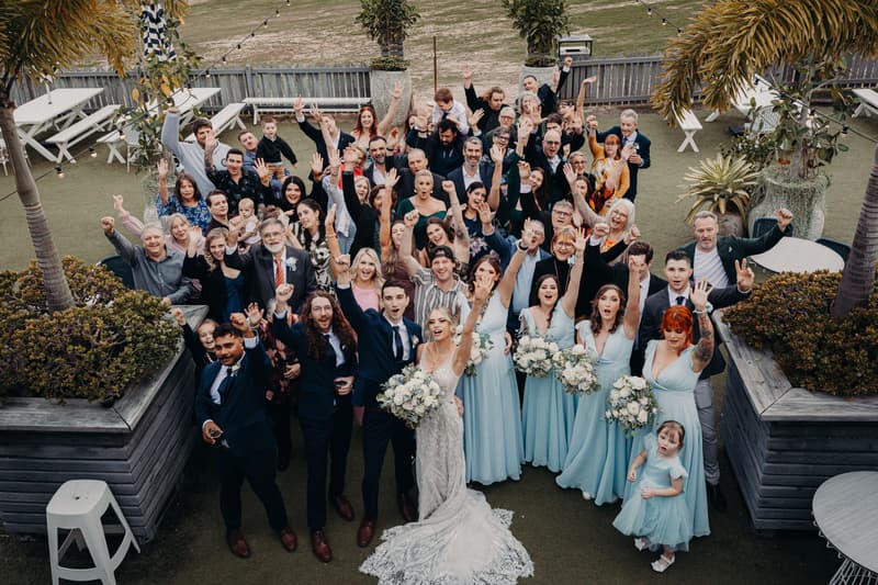 Courtney the bride and Cameron the groom pose with a large group of wedding guests and bridesmaids in light blue dresses at Sandstone Point Hotel, all raising their hands and cheering outdoors.